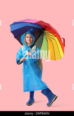 Graziosa studentessa in impermeabile con zaino e ombrello che cammina su sfondo rosa Foto Stock