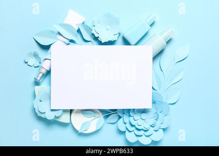 Set di prodotti cosmetici con cartoncino vuoto, fiori di carta e foglie su sfondo blu Foto Stock