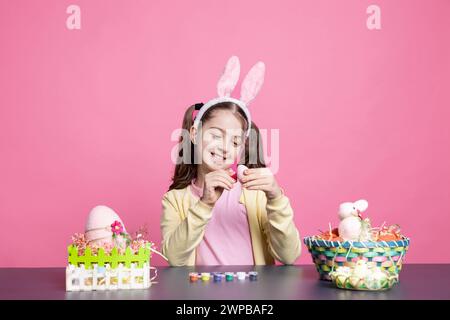 Un bambino adorabile che dipinge uova di pasqua con acquerello in studio, decorando ornamenti natalizi fatti a mano su sfondo rosa. I bambini più piccoli amano la creatività e l'artigianato, colorando con il pennello. Foto Stock