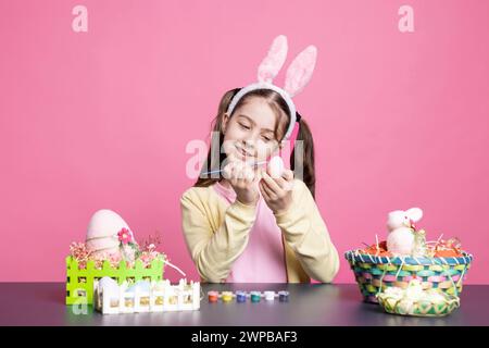 Dolce bambino che decora le uova di pasqua con tie dye sulla macchina fotografica, dipinge le decorazioni natalizie con acquerello su sfondo rosa. Il bambino più piccolo ama prepararsi e colorare con il pennello. Foto Stock