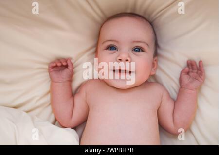 Un bambino appena nato sorridente e simpatico, che si stende su lenzuola beige nel letto o nella culla. Modello di imballaggio per prodotti per bambini. Bambino sano, concetto di ospedale e maternità felice. Neonato. Asilo nido. Foto Stock