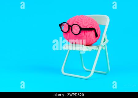 Modello di cervello umano rosa, decorato con occhiali, seduta su una sedia pieghevole verde su uno sfondo blu. Educazione, studio e impegno mentale verso conti Foto Stock