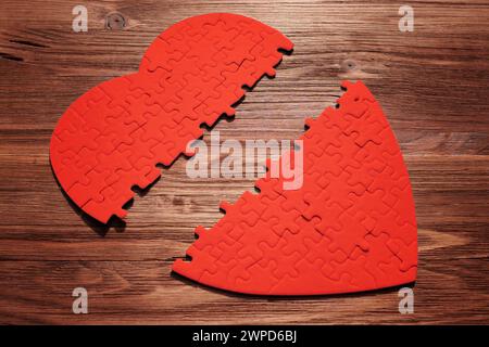 Puzzle rosso a forma di cuore, dimezzato e posato su una superficie di legno. I pezzi frammentati simboleggiano la disconnessione emotiva o le relazioni fratturate. Foto Stock