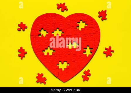 Puzzle rosso a forma di cuore poggiato su uno sfondo giallo, con pezzi mancanti disposti intorno ad esso. Sfondo creativo di San Valentino. Foto Stock