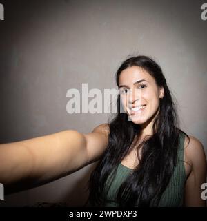 Una giovane donna allegra con lunghi capelli scuri cattura un momento felice con un selfie all'interno, mostrando la sua bellezza naturale e il suo sorriso contagioso Foto Stock