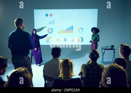 Uomo in piedi e che pone domande alle donne, agli esperti di business che presentano grafici di dati, con il pubblico che guarda. Foto Stock