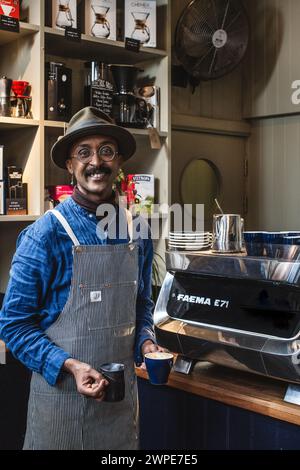 barista africano nero con grembiule per preparare caffè bianco piatto in caffetteria Foto Stock