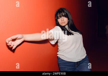 Ritratto di una giovane donna latinovenezuelana sdraiata sulla parete rossa, rilassata guardando la fotocamera, foto con luce dura. Foto Stock