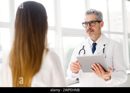 Un medico maturo con capelli grigi discute informazioni sulla salute su un tablet digitale con una giovane donna, in un ufficio clinico pieno di luce naturale Foto Stock