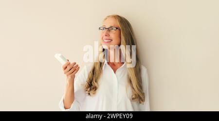 Ritratto di una donna di mezza età caucasica sorridente e felice accende il condizionatore d'aria o la TV utilizzando il telecomando a casa Foto Stock