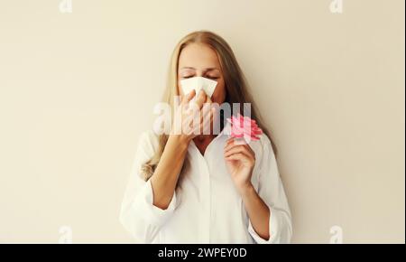 La donna malata e stanca, affetta da polline, tiene il naso soffiato con lo starnuti dei fiori utilizzando un tessuto su sfondo bianco dello studio Foto Stock