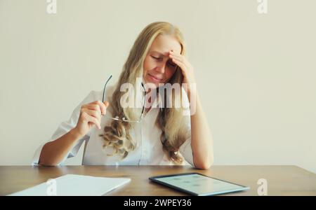 Lavoratrice di mezza età stanca e sovraccarica, affetta da mal di testa, si strofina gli occhi secchi, lavora con un tablet seduto alla scrivania in ufficio Foto Stock