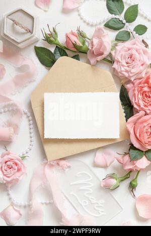 Carta e busta vuote vicino a rose rosa chiaro, anello di fidanzamento in una scatola e nastri di seta sul tavolo bianco, mockup di cartoleria per matrimoni. Romantico Foto Stock