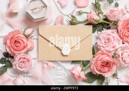 Busta sigillata in carta kraft vicino a rose rosa chiaro, anello di fidanzamento in una scatola e nastri di seta su un tavolo bianco, mockup nuziale. Appartamento romantico Foto Stock