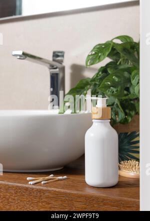 Un biberon bianco vicino al lavabo, monstera verde, spazzola per capelli di bambù e bastoncini di cotone su un ripiano in legno in bagno, primo piano, mockup cosmetico. Modalità Foto Stock