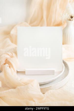 Tessera quadrata vicino al tessuto di tulle color crema sui piatti del tavolo della crema. Mockup di cancelleria per matrimoni. Arredi da tavolo con piccolo vaso e nodo in tessuto di seta, ro Foto Stock