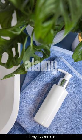 Bottiglia cosmetica bianca con un tiralatte su asciugamani piegati di colore blu vicino al lavandino e alla monstera verde, vista dall'alto, mockup. Scena Lifestile con prodotti di bellezza per la cura della pelle Foto Stock