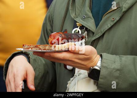 Parte centrale di un cliente, visitatore del mercato agricolo che tiene in mano un vassoio di carta con snack, fetta di pane con salsiccia grigliata. Primo piano, Long ang Foto Stock