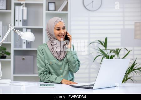 La giovane donna musulmana sorridente che indossa hijab ha avuto una piacevole conversazione sul suo smartphone al lavoro, con il laptop aperto. Foto Stock