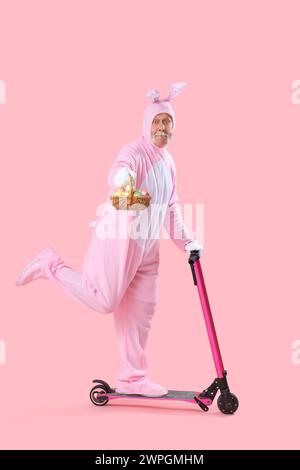 Uomo anziano in costume da coniglietto con uova di Pasqua in cestino di vimini che cavalcano scooter su sfondo rosa Foto Stock