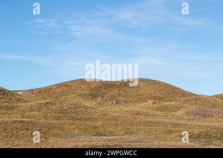 Tumulo sepolcrale ricoperto di erba in un paesaggio di cultura collinare ondulato. Macchie di neve a terra e cielo limpido con nuvole leggere Foto Stock