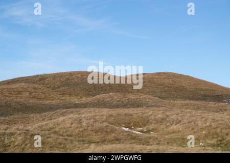 Tumulo sepolcrale ricoperto di erba in un paesaggio di cultura collinare ondulato. Macchie di neve a terra e cielo limpido con nuvole leggere Foto Stock