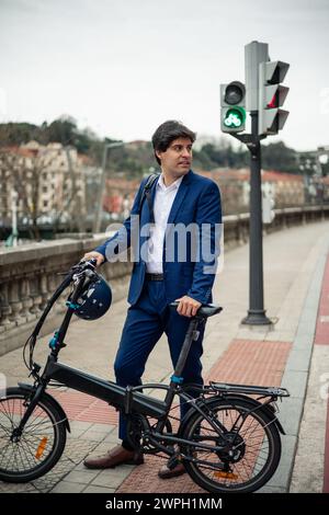 Ritratto verticale Un giovane uomo d'affari posa con sicurezza tenendo la sua bici elettrica pieghevole e il casco in un ambiente urbano. Questa immagine riflette la sua comunicazione Foto Stock