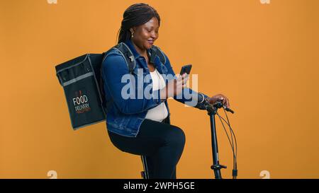 Donna afro-americana multitasking sulla sua bici, che consegna cibo con il cellulare in mano. Giovane donna nera che controlla l'app di consegna su dispositivo mobile per un trasporto efficiente degli ordini. Foto Stock
