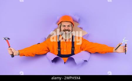 Costruttore sorridente in elmetto con chiave regolabile e martello che guarda attraverso il foro della carta. Uomo barbuto in uniforme da costruzione e casco protettivo Foto Stock