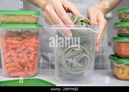 Donna che mette la pasta cruda in un contenitore di plastica al tavolo leggero in cucina, primo piano. Conservazione degli alimenti Foto Stock