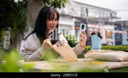 Una donna asiatica allegra ed eccitata guarda lo schermo del suo smartphone con un volto estremamente felice, ha ricevuto una buona notizia inaspettata, seduta a un tavolo Foto Stock