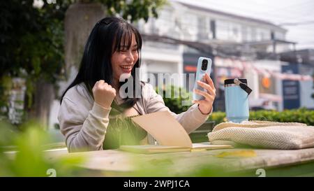 Una donna asiatica allegra ed eccitata guarda lo schermo del suo smartphone con un volto estremamente felice, ha ricevuto una buona notizia inaspettata, seduta a un tavolo Foto Stock