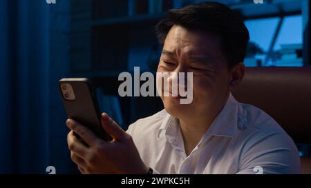 Uomo adulto asiatico di notte in ufficio di casa perso fallimento con il telefono cellulare triste infelice cinese maschio con smartphone perdere cattive notizie frustrato turbato Foto Stock