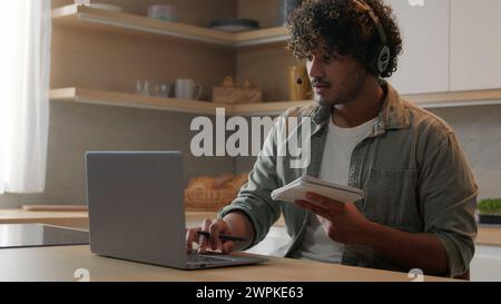 Indian man in headset look app pc browsing typing laptop write ideas questions notes review research sit at home kitchen. Arabian guy learn language Stock Photo