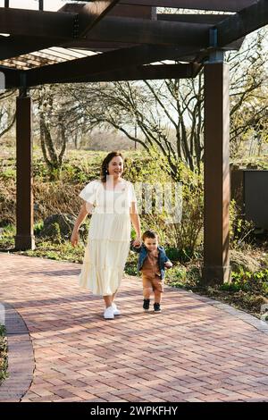 La mamma cammina lungo il sentiero in mattoni tenendo il figlio in mano in giardino Foto Stock