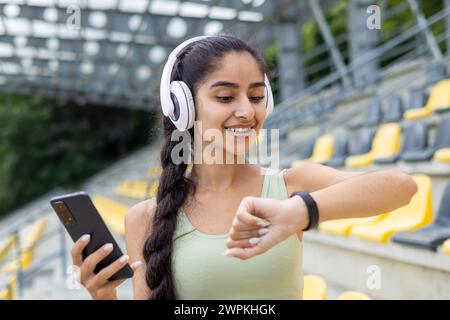 Una donna indiana in abbigliamento sportivo controlla il suo fitness tracker durante una sessione di jogging in uno stadio con le cuffie indossate. Foto Stock