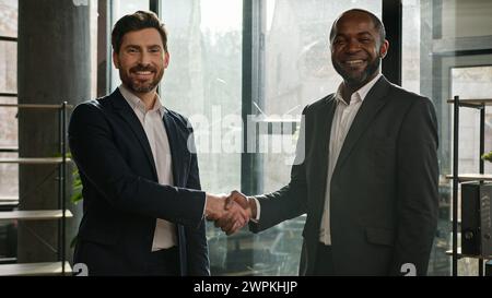 Due diversi uomini d'affari multirazziali colleghi d'affari partner uomini che stringono in ufficio posa sorridenti a cinepresa uomo caucasico e afroamericano Foto Stock
