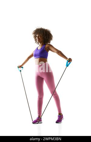 Felice donna afroamericana che si allena con bande di resistenza, vestita con abbigliamento sportivo viola-rosa su sfondo bianco studio. Foto Stock