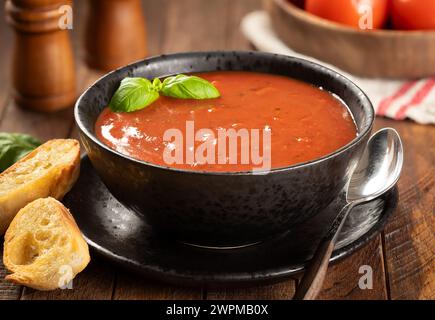 Ciotola di zuppa di pomodoro guarnita con foglie di basilico e fette di baguette tostate su un tavolo di legno Foto Stock