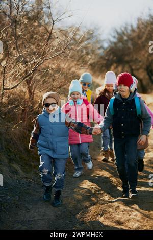 I bambini felici e divertenti che si tengono per mano indossando abiti invernali e zaini vanno sulle piste soleggiate. Attività all'aperto per lo sviluppo dei bambini. Foto Stock