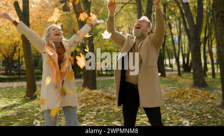 Felice spensierato vecchio caucasico coppia amore donna in pensione e uomo che lanciano foglie gialle nella foresta autunnale del parco insieme godere di caduta foglie divertente maturo Foto Stock