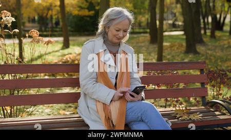 Felice dai capelli grigi bella donna matura che usa lo smartphone online nel parco autunnale donna caucasica che utilizza tecnologie Internet al di fuori della signora anziana Foto Stock