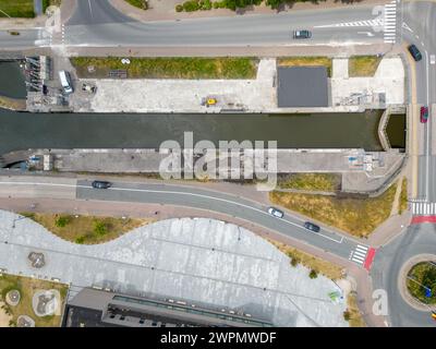 La vista a volo d'uccello in questa fotografia cattura l'intersezione delle infrastrutture urbane dove convergono le strade e un corso d'acqua. La composizione è uno studio di geometria e organizzazione, che mostra il layout pianificato della città. I veicoli appaiono simili a giocattoli da questa altezza, muovendosi lungo le corsie segnalate, mentre il corso d'acqua corre parallelo, delimitato da bordi di cemento. Il cavalcavia crea un ulteriore strato del paesaggio urbano, dimostrando la complessità delle reti di trasporto in una città moderna. L'immagine funge da esempio di pianificazione urbana, evidenziando le diverse modalità di trasporto Foto Stock
