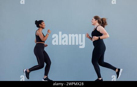 Donna sportiva che corre di fronte alla sua amica e che è motivata a correre. Due amici di jogging sorridono mentre corrono al muro grigio. Foto Stock