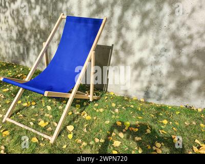 Una sedia a sdraio blu si erge alla luce del sole su un prato verde davanti a una parete di cemento grigio chiaro con ombre di foglie Foto Stock