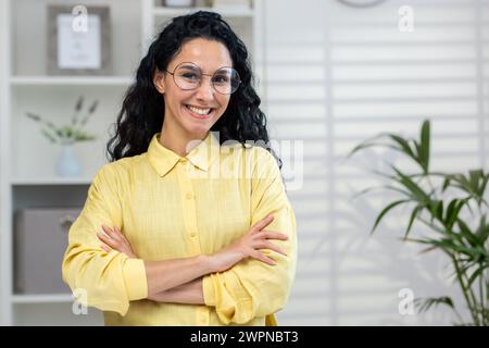 Una donna professionista con gli occhiali è in piedi con sicurezza, le braccia incrociate, in un ambiente d'ufficio ben illuminato, per conferire potere e positività. Foto Stock