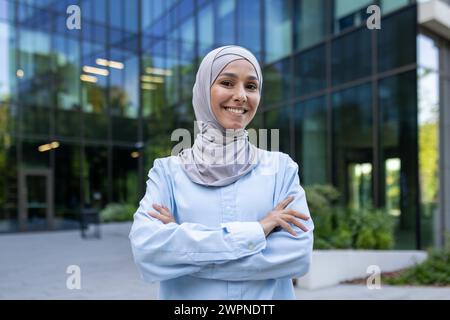 Giovane donna musulmana professionista con un sorriso irresistibile, con indosso un hijab, in piedi con sicurezza fuori da un ufficio. Foto Stock