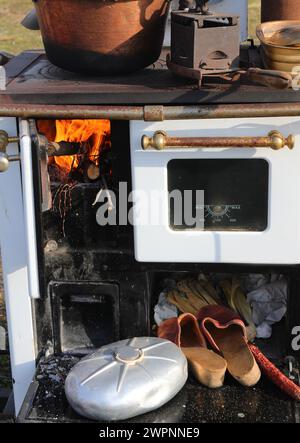 Vecchia stufa a legna in una cucina di fattoria con un fuoco acceso e una pentola per riscaldare l'acqua e vecchie scarpe contadine con un calderone in rame sulla parte superiore Foto Stock
