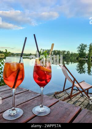 Due bicchieri di Aperol Spritz con fetta di paglia e limone su un tavolo, molo di legno vicino al lago con sdraio e vista sull'acqua, cielo blu con clou Foto Stock