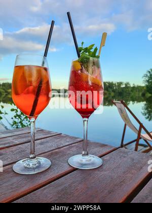 Due bicchieri di Aperol Spritz con fetta di paglia e limone su un tavolo, molo di legno vicino al lago con sdraio e vista sull'acqua, cielo blu con clou Foto Stock